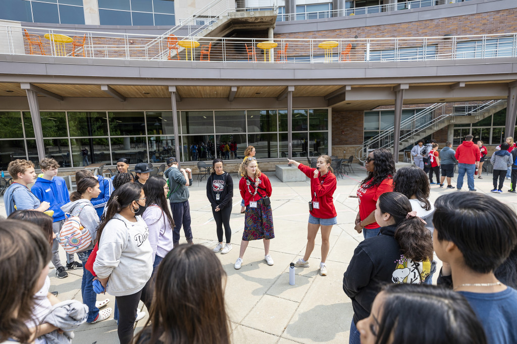 Students stand around a plaza in a circle, listening to a woman talk.