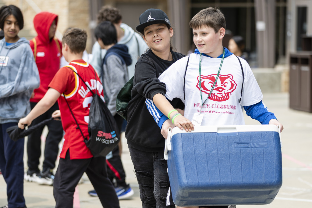 Some kids walk and carry a cooler.