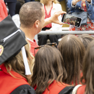 A group of people, including one wearing commencement robes, gather together as one of them takes a selfie photo with a cell phone.