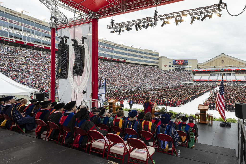 A picture taken from the back of the stage, showing stands filled with graduates and their family and friends.