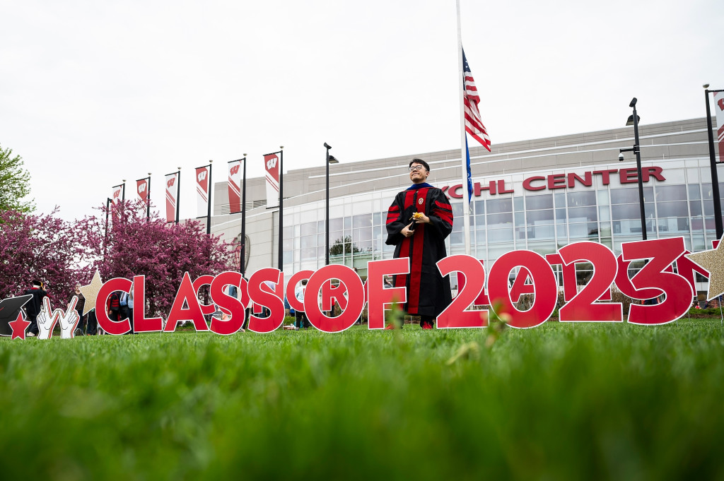 A person poses in front a sign on a lawn, with an arena in the background.