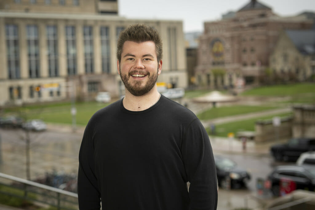 A portrait photo of Liam McLean standing outside with a green lawn and campus buildings out of focus behind him.