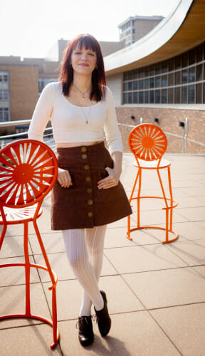 A photo of Meg Mercy standing next to a metal sunburst chair outdoors at Union South.