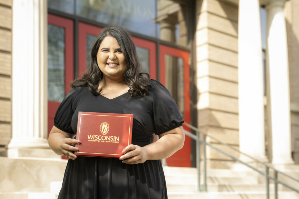 Yasmeena Ougayour stands outside the School of Education, a sandstone building with white columns and red doors. She is smiling to the camera and holding a red diploma folio with the University of Wisconsin–Madison seal and the words "Wisconsin University of Wisconsin–Madison."