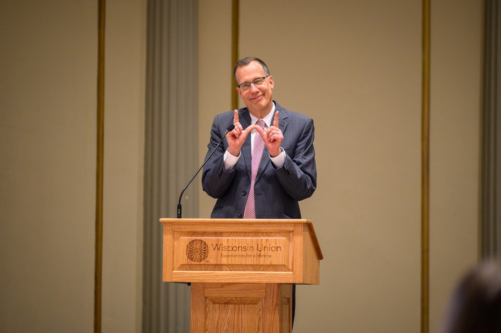 Standing at a lectern, Karl Scholz makes a W sign with his thumbs and forefingers.