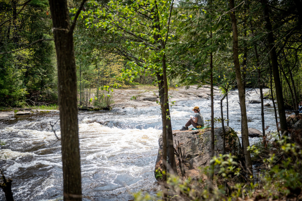 A woman sits on a rock by a rushing river.