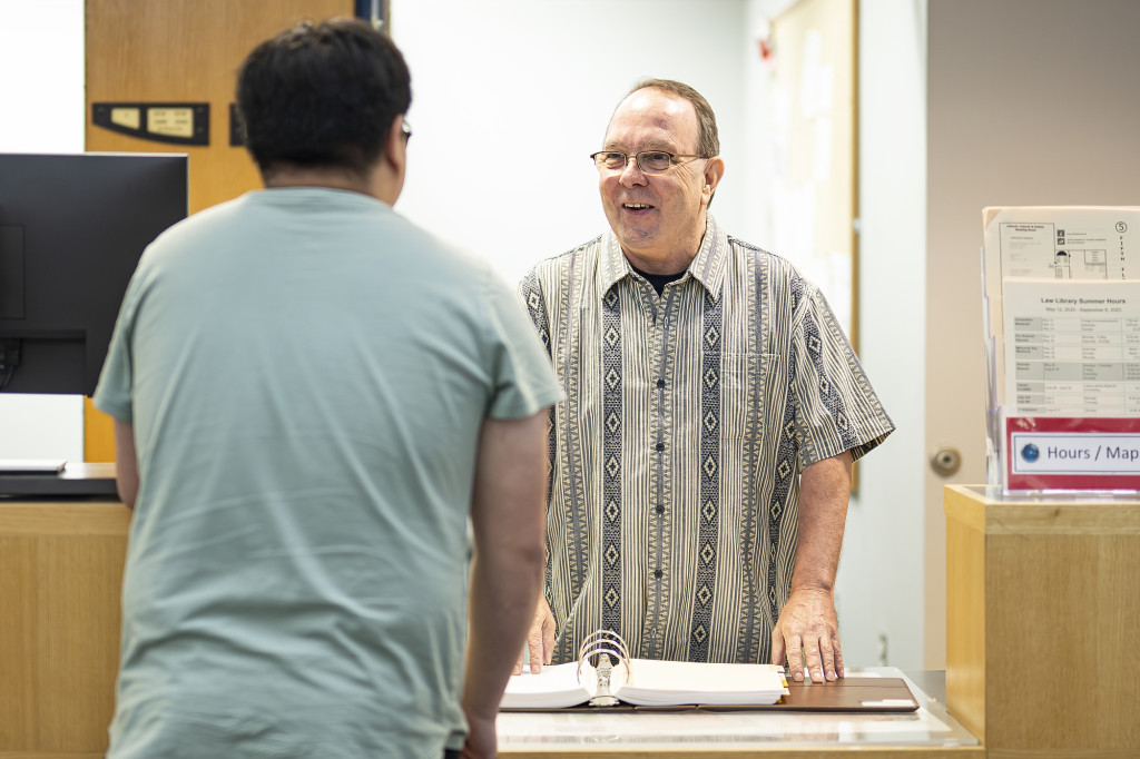 A man at a desk stands and talks to another man, whose back we see.