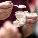 A close up shot of a person's hands holding a cup of ice cream and scooping a bite with a spoon.