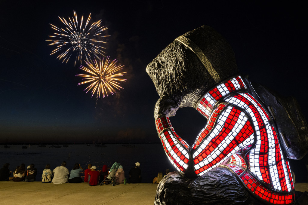 A statue of Bucky Badger is posed sitting with his cheek resting on his hand. He is looking away toward Lake Mendota where a fireworks display lights the sky.