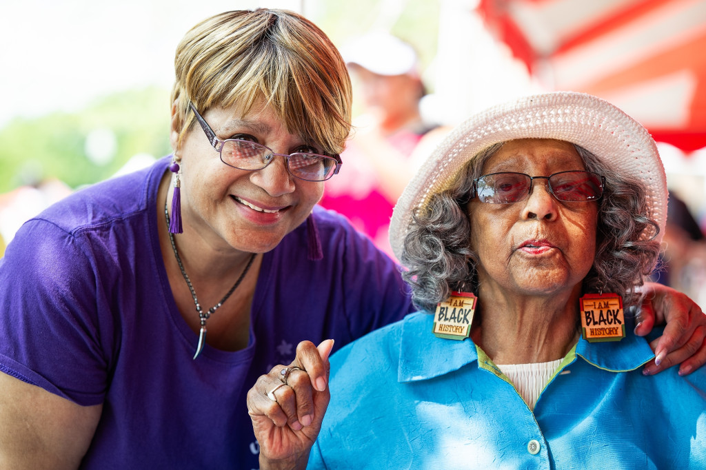 Two women lean together and smile to the camera. Sarah Wells, seated on the right, wears earrings that say I am Black History.