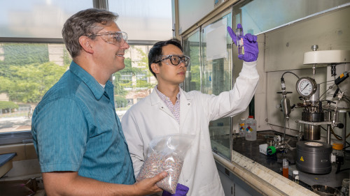 Two men stand in a chemistry lab wearing safety goggles. One, wearing a blue collared shirt, stands to the left holding a bag full of shredded plastic. The other, wearing a white lab coat and purple gloves), holds a vial up to the light for both to examine.