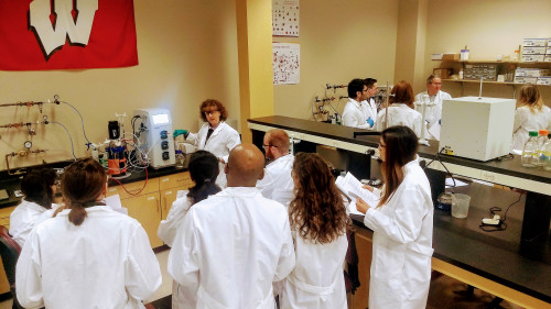 In a laboratory, people in white coats gather around a woman operating a piece of equipment. A UW–Madison flag hangs on the wall.