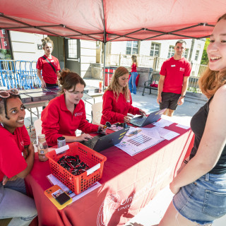 Three people in red shirts sit at a table under a red tent. They're talking with students who've come up to the table.