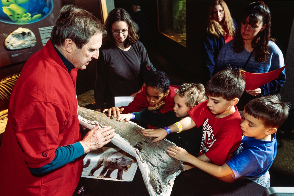 A man talks about a dinosaur bone from a triceratops. Children listening to him, reach out to touch the bone.