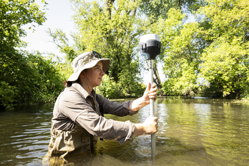 A woman wades in a creek with equpment.