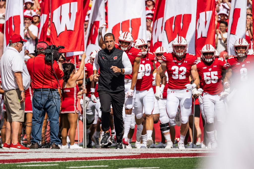 A man in black pants and sweater vest jogs onto the field with players in red football uniforms.