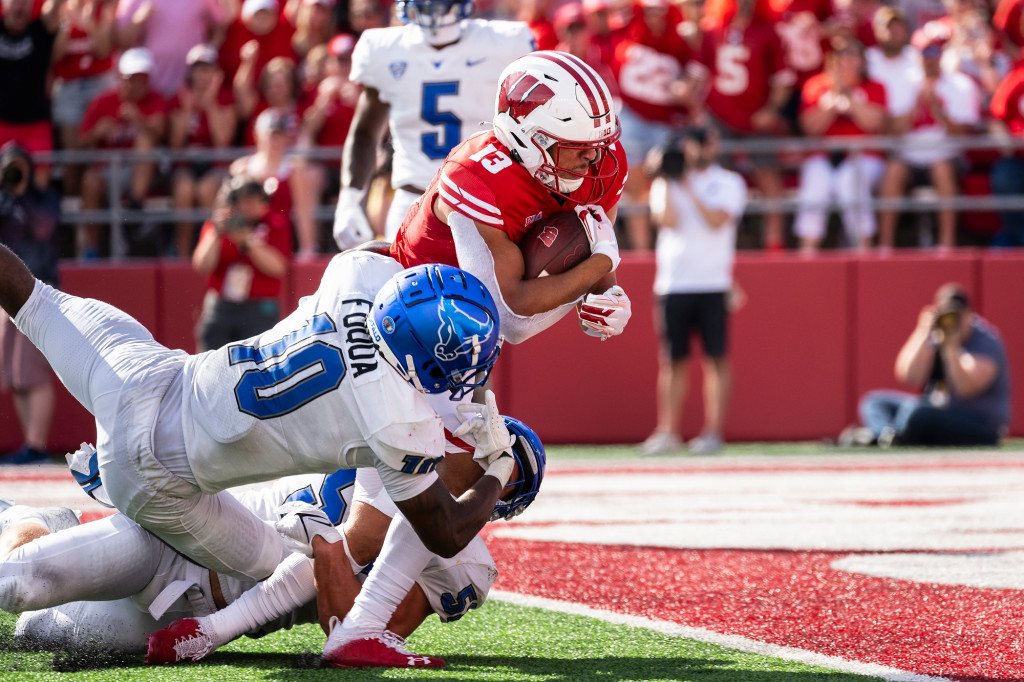 A man in a red football uniform dives into the end zone as another tries to hold him back.