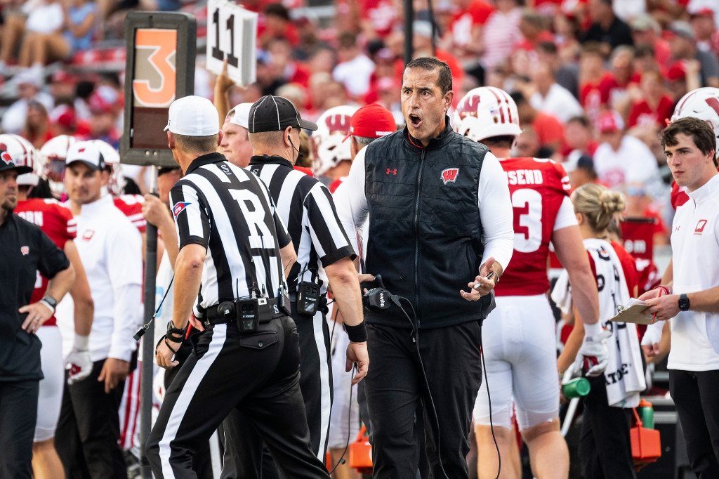 A man on the sidelines talks with referees in striped shirts.