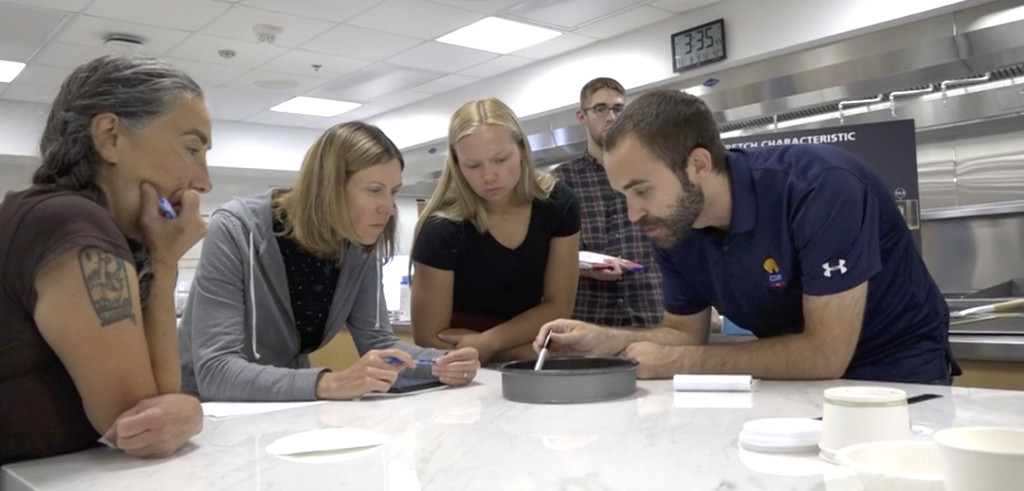 Four people in an industrial kitchen space lean over a table and look into a round metal pan.