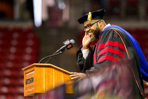 LaVar Charleston, wearing academic robes, stands at a lectern. He's holding one hand to his ear and smiling as he listens to the crowd.