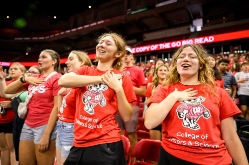 A group of four students in a crowd close their eyes, hold one hand to their hearts and sing along to a song.