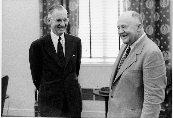 In this black and white photo, two men wearing suits and ties stand in a formal office setting.