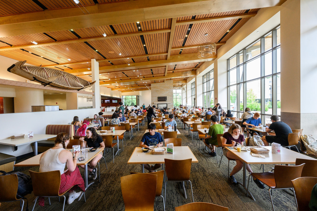 Students sit at tables in a cafeteria eating, with large windows facing them.