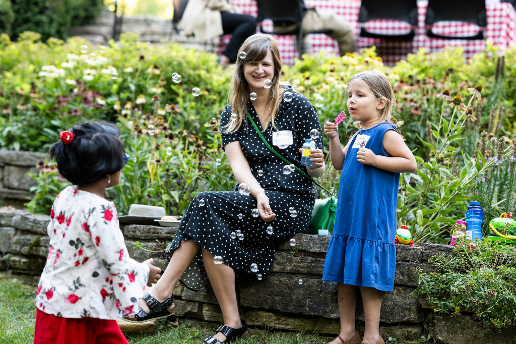 A young girl blows bubbles to another young girl on the lawn of Olin House. A woman sits on a low stone wall and smiles as she watches the girls.