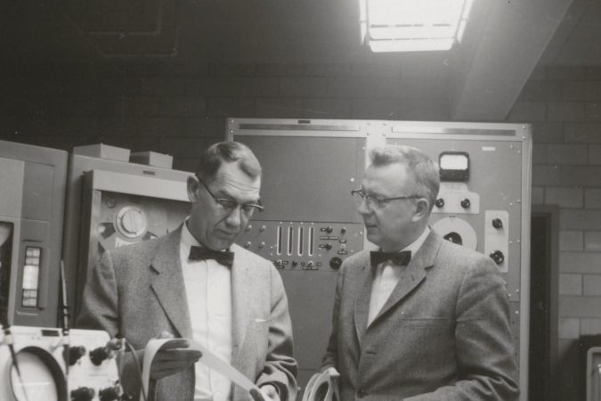 In a black and white photo, two men wearing suits and bow ties read from a ticker tape printed from a large, room-sized computer behind them.