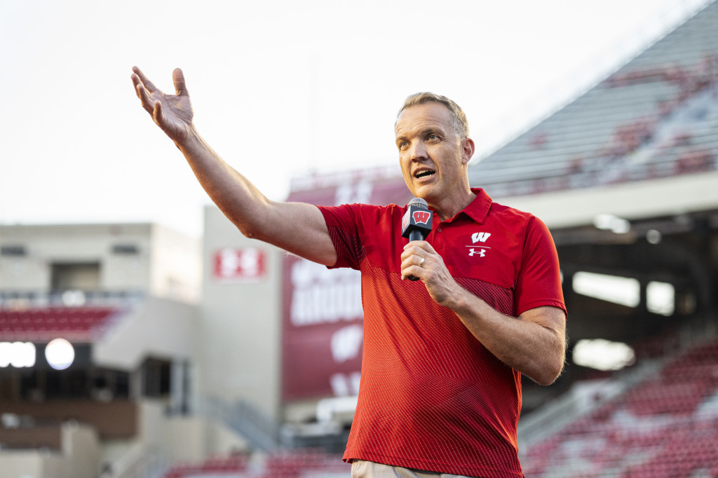 Chris McIntosh speaks to the crowd. He's holding a microphone in one hand while he gestures with the other.