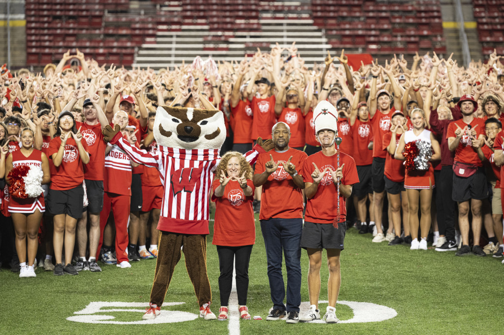 Bucky Badger, Chancellor Mnookin, Provost Isbell and a marching band marshall smile and make W signs in front of the W at the 50 yard line.