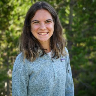 Lucy McGuire smiles in front of a stand of lush, green lodgepole pines.