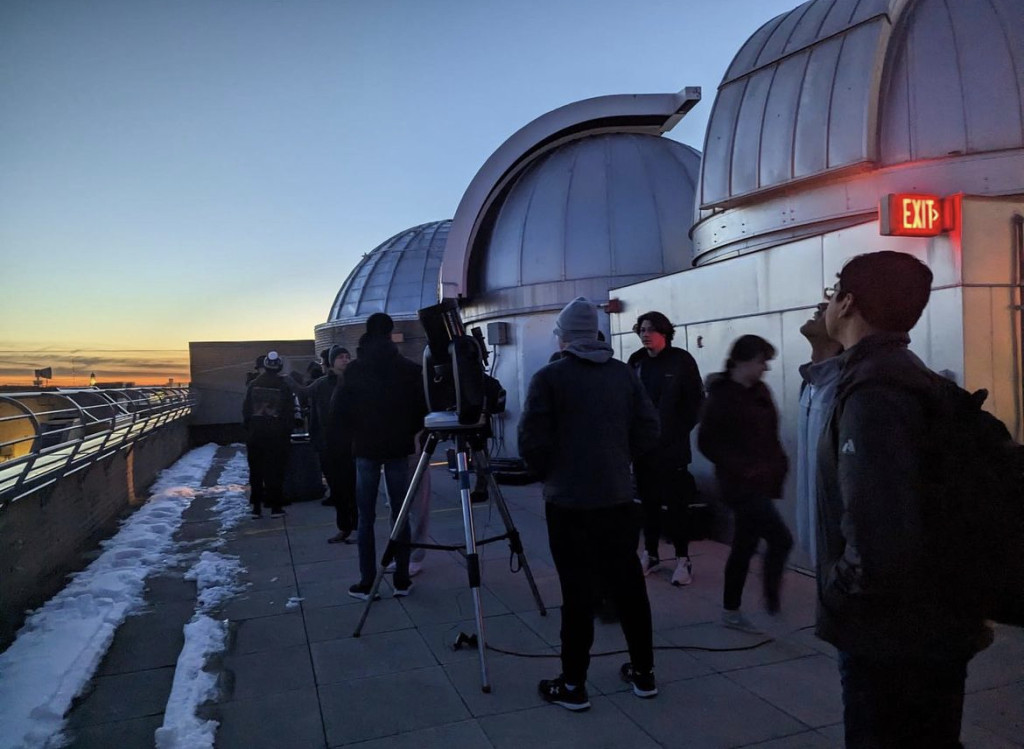 A group of people wearing winter clothes gather on a rooftop with snow visible.