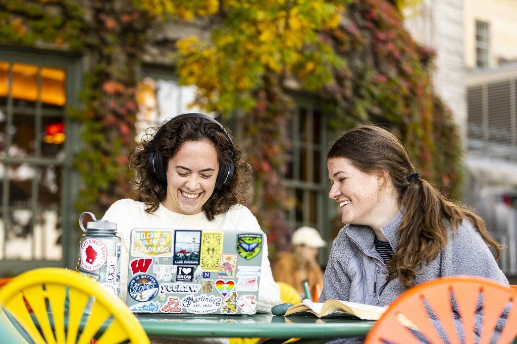 Two friends laugh together as they sit outside at a green metal table with yellow and orange chairs. The ivy growing on the wall behind them has changed from green to yellow and red.