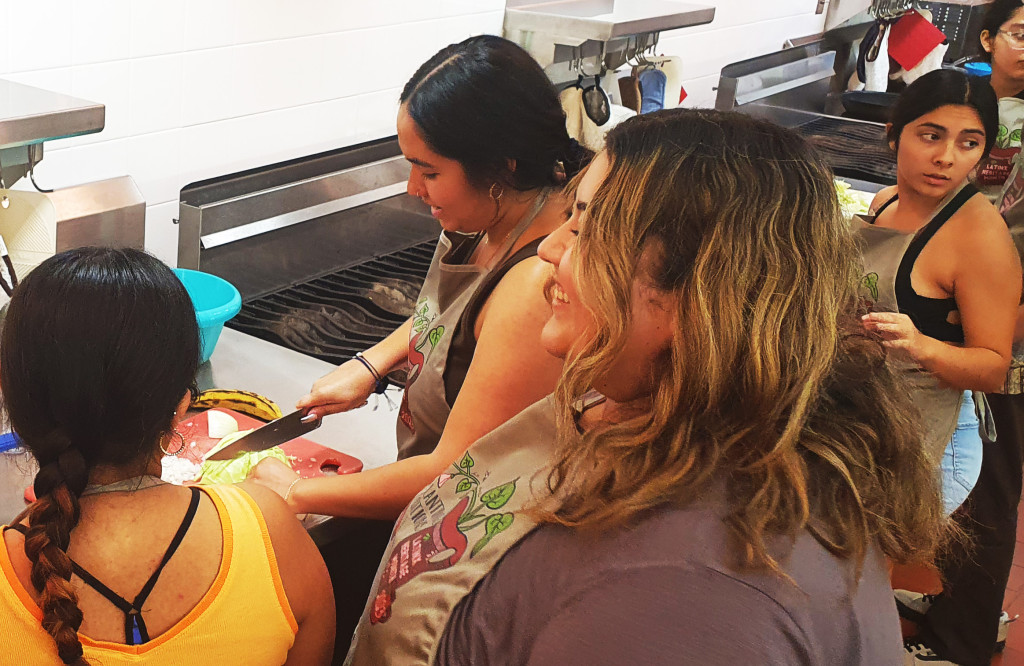 A woman stands at a cutting board chopping an onion while two friends watch from behind.