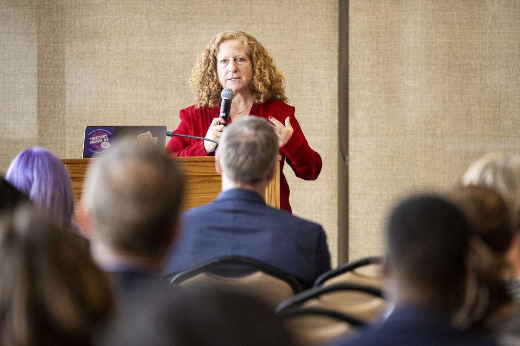 A woman speaks from a podium to a seated audience.