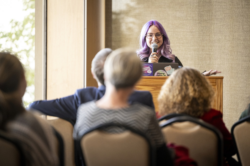 A woman speaks from a podium to a seated audience.