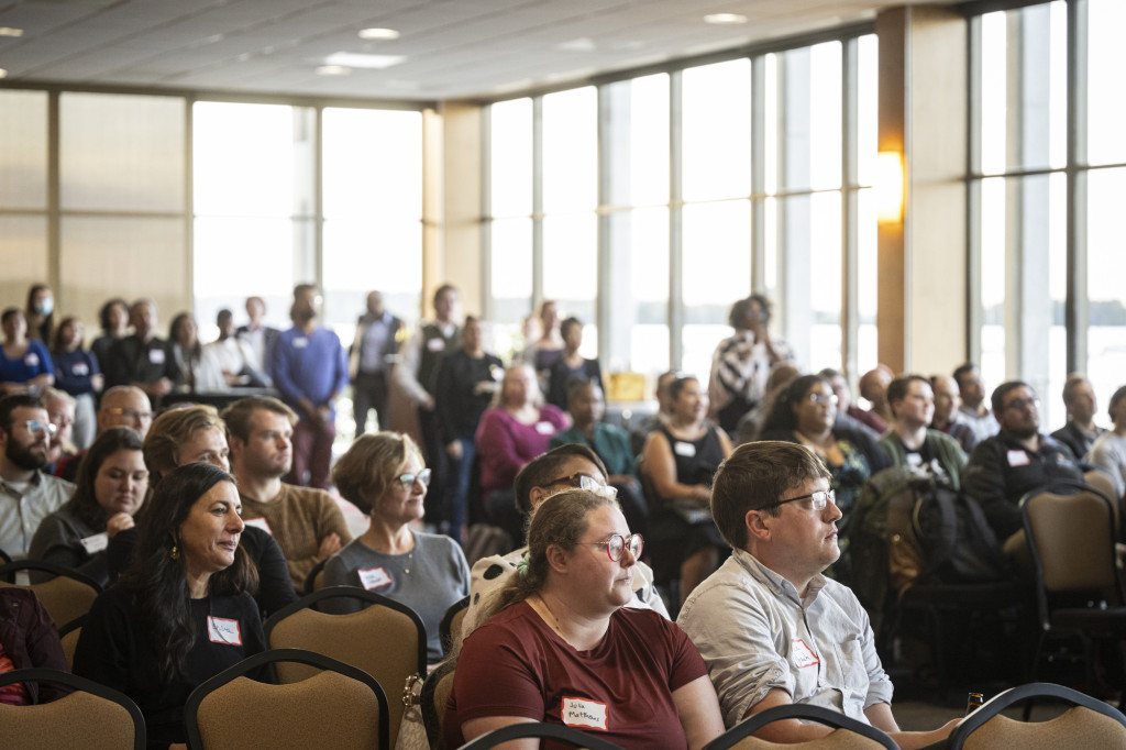 People sit in rows listening to an unseen speaker in a bright room with floor-to-ceiling windows.