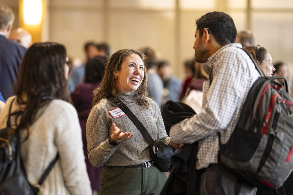 Amidst a crowded room, three people stand in conversation with one another.