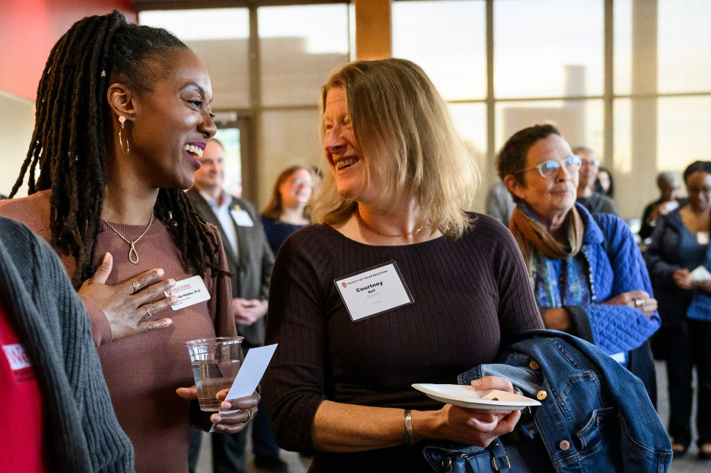 Two women in a crowded reception room stand side by side, smiling and laughing.