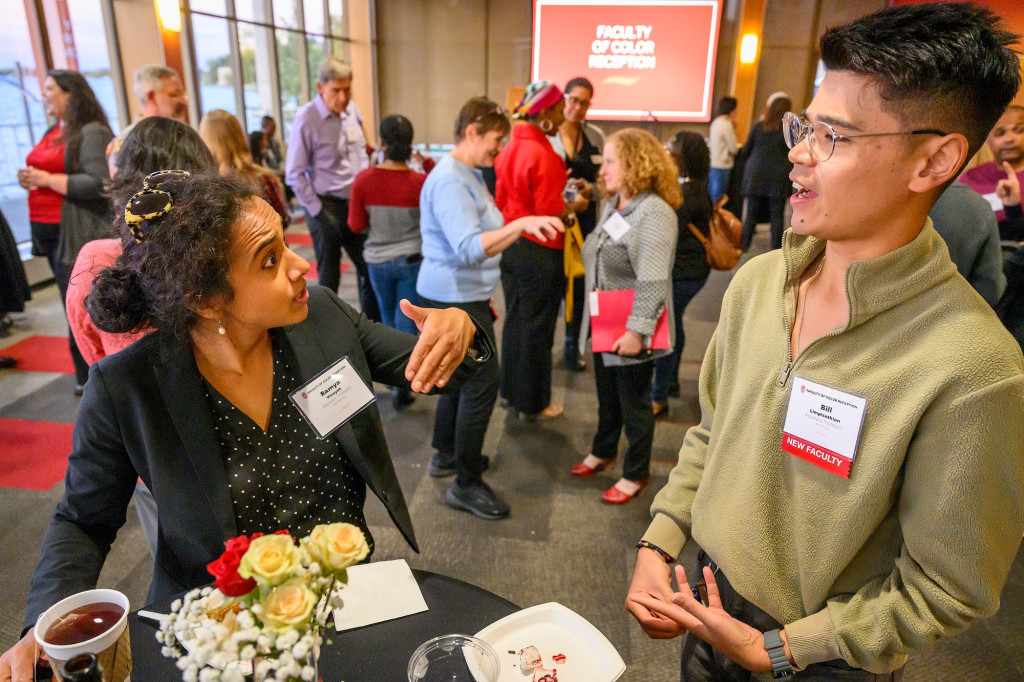 In a crowded reception room, two people stand at a cocktail table and speak with one another.