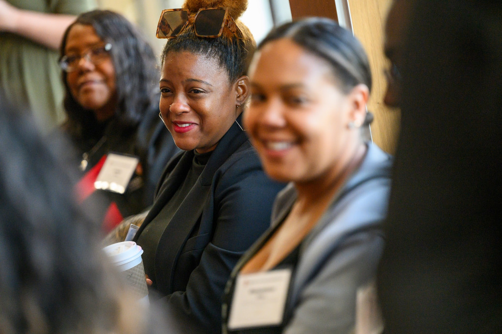 Three women seated at a reception smile as they listen to an off-camera speaker.