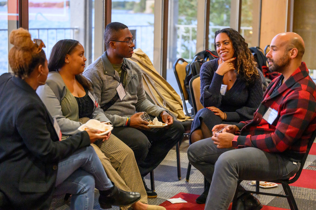 A group of five people in conversation sit in chairs in a semi circle in a crowded reception room.