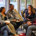 A group of five people in conversation sit in chairs in a semi circle in a crowded reception room.