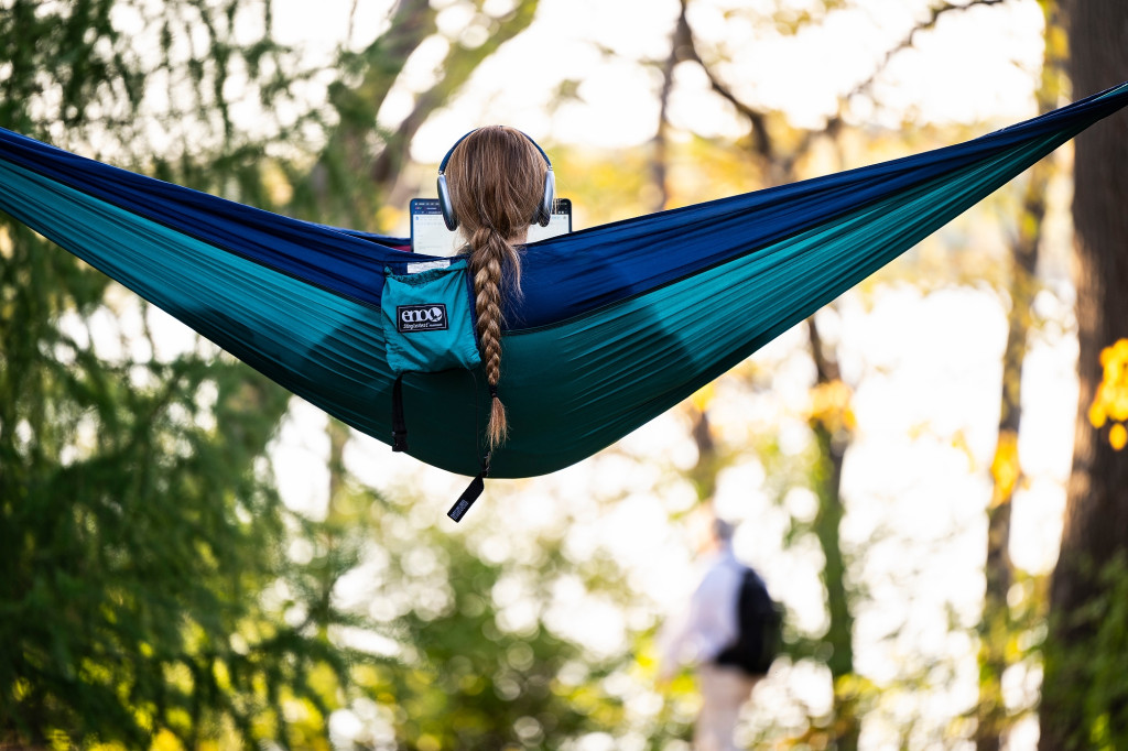 Facing away from the camera, a person with a long braided ponytail sits in a blue hammock. They are wearing over-ear headphones and looking at a laptop.