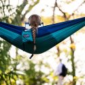 Facing away from the camera, a person with a long braided ponytail sits in a blue hammock. They are wearing over-ear headphones and looking at a laptop.