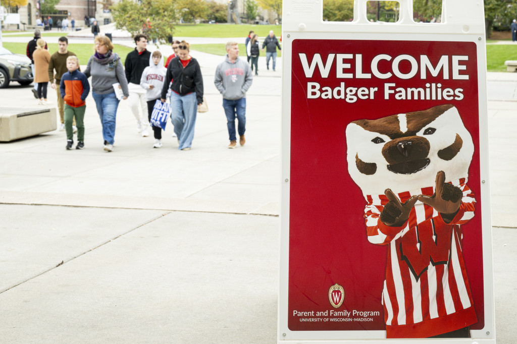 A group of people walk on a sidewalk, a sign in the foreground reads 