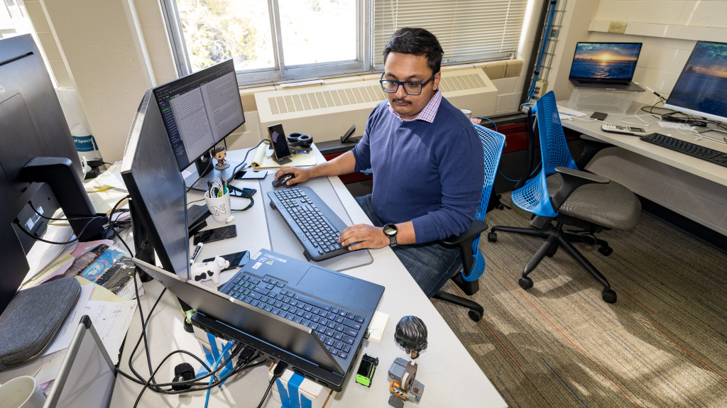 A mab sits at a desk with two monitors and a laptop. He's typing on an external keyboard while looking at the laptop screen.