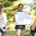 A man holds up a large map showing the locations of effigy mounds on the UW campus. A woman standing next to him looks on as he speaks to a gathered crowd (not seen).