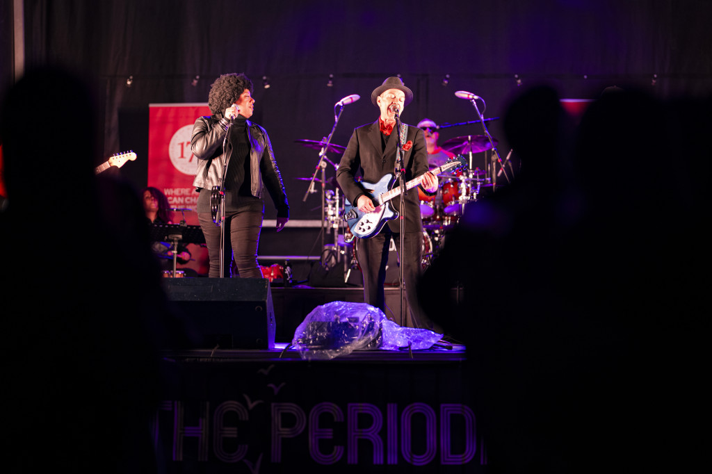 In a photo of a rock band on stage, a woman at a microphone looks to her bandmate as he sings and plays guitar.